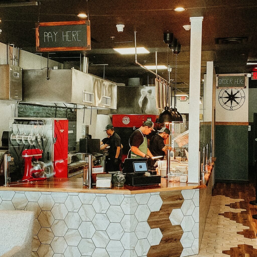 Interior of a modern restaurant kitchen with staff preparing food. The counter has hexagonal tiles, and overhead signs indicate areas for payment and ordering.