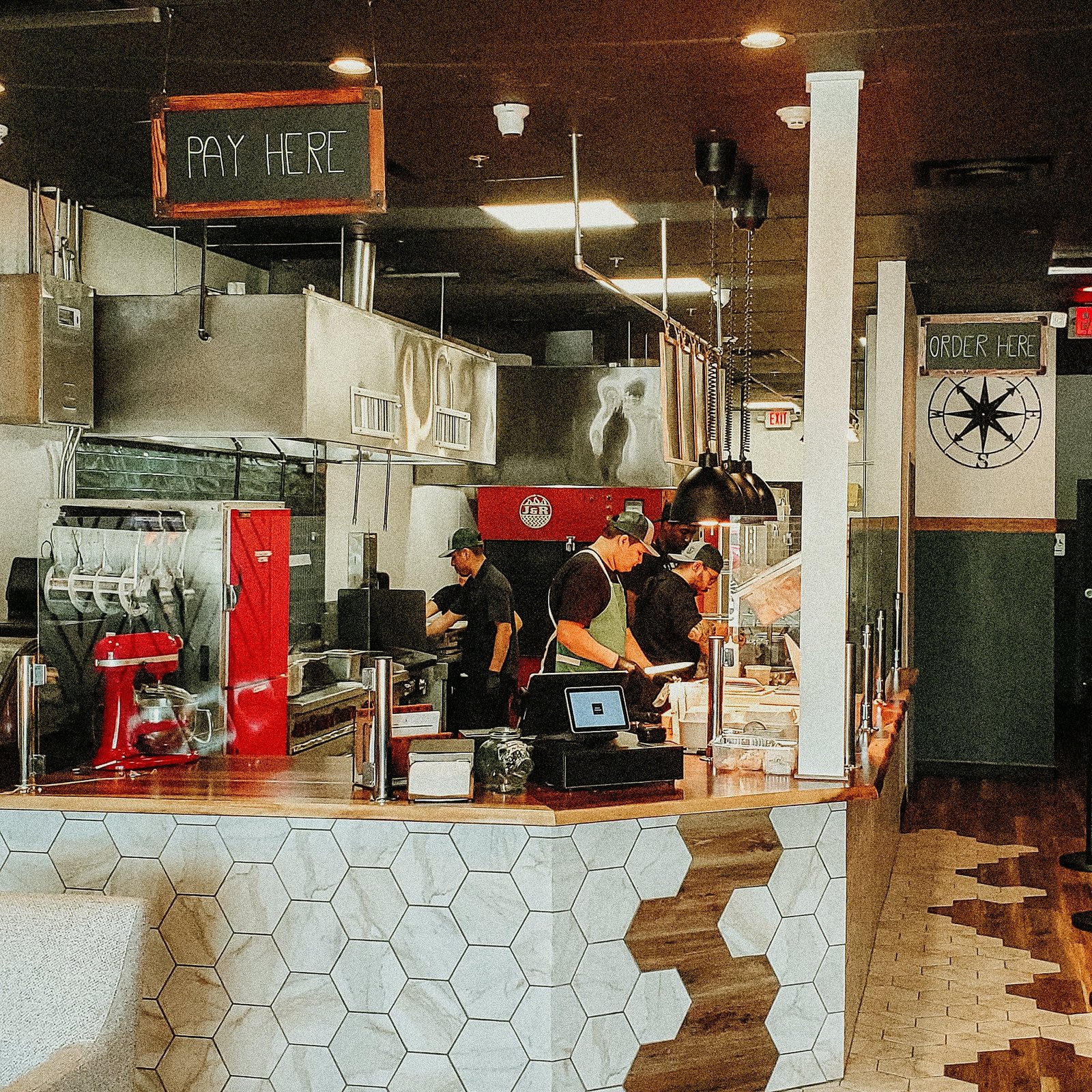 Interior of a modern restaurant kitchen with staff preparing food. The counter has hexagonal tiles, and overhead signs indicate areas for payment and ordering.
