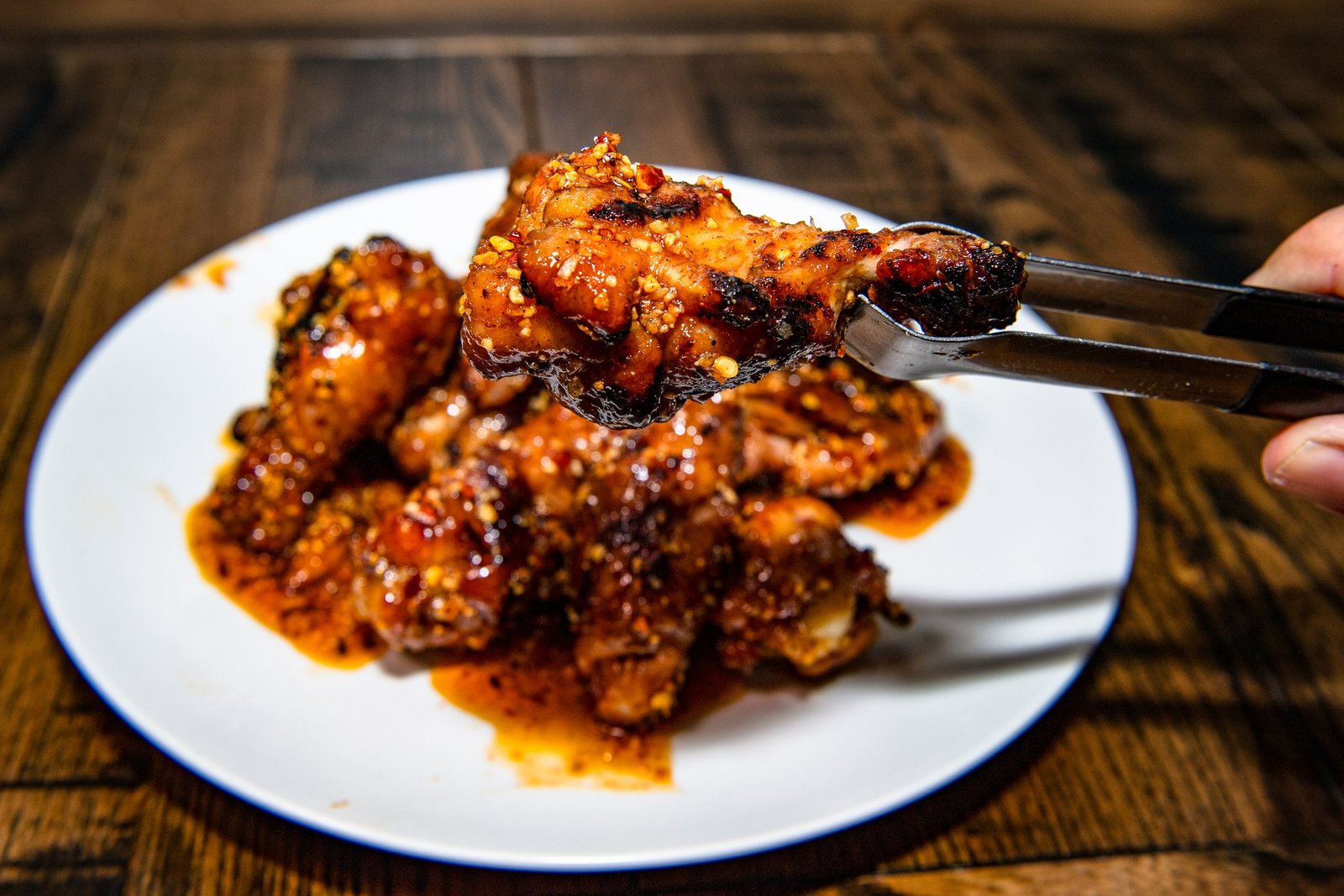 Close-up of sauced chicken wings on a white plate, with a hand holding tongs in the foreground.