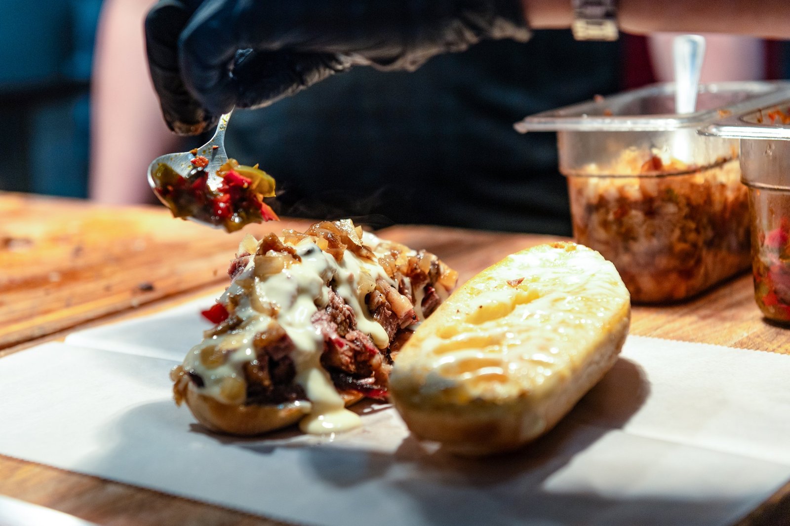 Person wearing black gloves serving loaded baked potato with sour cream, cheese, bacon, and vegetables on a white paper-lined wooden surface, with containers of toppings in the background.